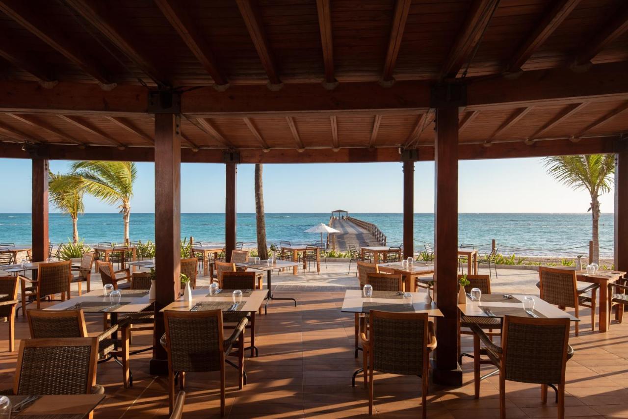 Impressive Punta Cana Exterior foto The photo shows a beachfront dining area with a wooden structure featuring a thatched roof. There are several tables and chairs arranged on a wooden deck, providing a picturesque view of the ocean. Palm trees can be seen swaying in the breeze, and th